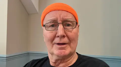 Steve Jones/BBC Derek Wilson, un hombre mayor que lleva una diadema naranja. También lleva gafas y mira directamente a la cámara.