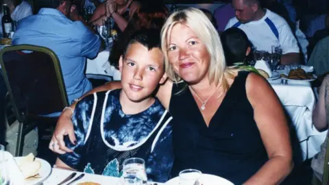 Matthew Leahy da ragazzo con sua madre Melanie. Sono seduti a un tavolo da pranzo con altri commensali sullo sfondo. Melanie ha abbracciato suo figlio, che è seduto alla sua destra. Matthew indossa una maglietta da basket sopra una maglietta tie-dye e ha i capelli corti e neri. Melanie indossa un top nero e una collana e ha i capelli biondi di media lunghezza.