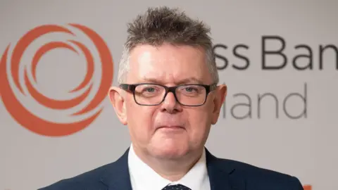 Brass Band England A man with short grey hair and glasses wearing a suit standing in front of an orange and white Brass Band England logo.