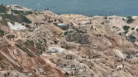 Overhead shot of mining activities at Rubaya mine where the terrain is hilly - slightly terraced. The bare earth is a light brown, some of it has a darker more orange shade. Many groups of miners can be seen from a distance cleaning ore. One corrugated metal shack can be seen. In the distance are green fields.
