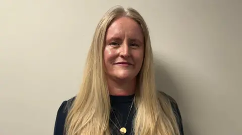 Geraldine Hanna - a woman with long blonde hair is stood in front of a plain, white wall looking at the camera. She is wearing a dark, long sleeve top with a gold necklace.