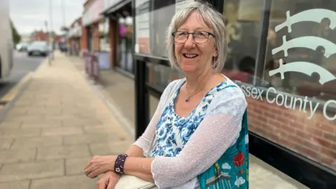 Stuart Woodward/BBC Pat Reeves sits at a bus stop in Hockley with a row of shops behind her. She is wearing a blue and white floral top with a white cardigan over the top. She has a colourful bag over her shoulder, and has grey hair and wears glasses.