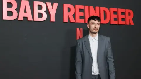 Getty Images Richard Gadd, looking thin and pale in a white shirt and grey suit, in front of a grey wall and the name Baby Reindeer logo in red lettering.