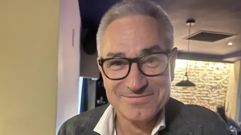 Gerald Voisin is pictured wearing a grey blazer, a white shirt and black-framed glasses. He has light grey hair and is smiling at the camera.