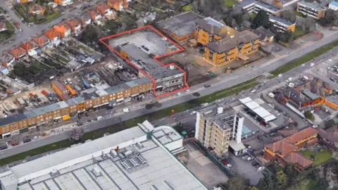 Bond Wolfe An aerial image of the site, which is marked out by an orange line. Other buildings are to the left and right of the site on the road.