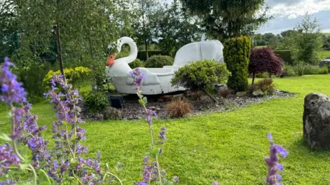 A swan boat on a platform in a garden, surrounded by buses and bark. Grass circles the boat and lilac flowers, bushes and other trees can be seen.