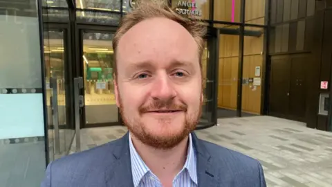 Adam Brown, with short brown hair, wearing a blue jacket and blue and white striped shirt, standing outside an office building with rotating glass doors.