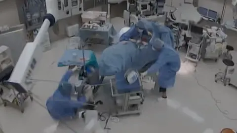 CCTV image of an operating room with doctor falling over and grabbing the operating table