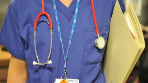 A close up image of a doctor in scrubs, they are wearing a stethoscope and carrying a file. 