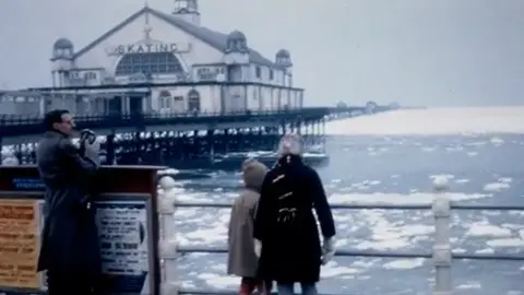 John Clague Two children and a man with a camera stand at the railings on the promenade looking out onto a frozen sea. The skating rink on Herne Bay pier can be seen in the background. 