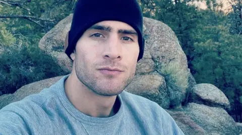 Keith Milkie Keith Milkie, a young man wearing a grey sweatshirt and a dark beanie, pictured on a rocky outcrop with a forest behind him