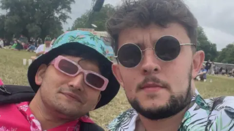Ashwin and Connor take a selfie while sitting in a field during a festival. They both wears sunglasses and floral shirts. 