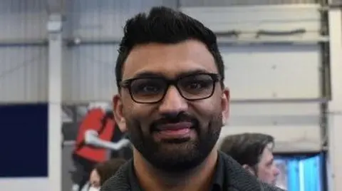 Imran Latif with dark hair, beard and moustache, wearing black glasses and a black jacket