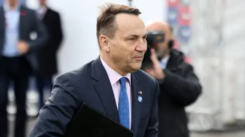 Menteri Luar Negeri EPA Polandia Radoslaw Sikorski berjalan sambil memegang map kulit hitam. Dia mengenakan blazer biru tua, kemeja merah muda muda, dan dasi kotak-kotak biru. Dia memiliki rambut coklat pendek dan mata biru.