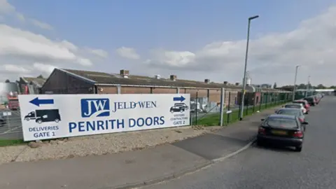 The outside of the JELD-WEN building. There is a large white sign saying JELD-WEN PENRITH DOORS on a green metal fence. A single-storey factory can be seen in the background. Cars are parked nearby.