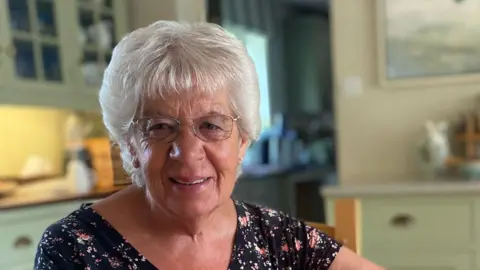 Doreen Savage smiles at the camera. She has short grey hair and wears a black floral top. She also wears glasses.