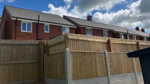 A pale wooden fence in the foreground can be seen with a row of red brick houses behind it. The windows of the upper floor of the houses are visible above the level of the fence.