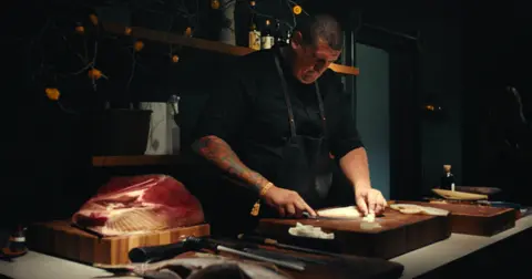 Aberystwyth Town Football Club Ward cutting fish on a brown wooden chopping board. He is wearing a black top and a dark apron on top and has a sleeve tattoo on his right arm. There is a slab of red meat on the side too. 