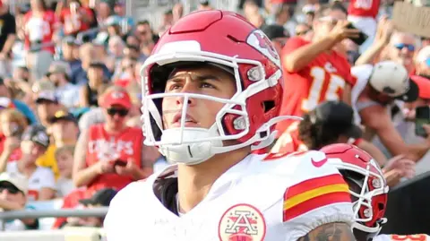 Getty Images Louis Rees-Zammit enters the field in the red and white kit of the Kansas City Chiefs. A red number 9 is on his white shirt and he wears a red American football helmet. Louis wears shoulder pads and has a heavily tattooed right arm. A crowd dressed in red cheers behind him. 