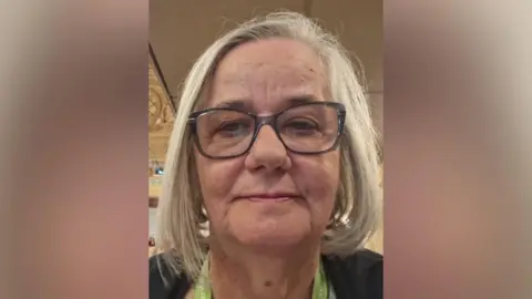A close-up portrait of Helen Edwards with shoulder-length grey hair wearing dark-framed glasses, photographed indoors against a softly lit background.
