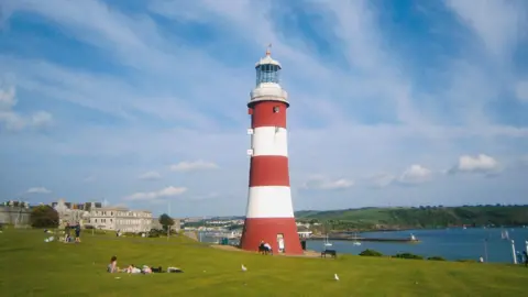 Smeaton's Tower, the lighthouse on Plymouth Hoe, on a sunny day with a blue sky and puffy white clouds. The lighthouse is white with three broad brick-red stripes around it. In the foreground, a handful of people are lying on the green slopes of The Hoe with a few seagulls strutting about while Plymouth Sound (the body of water forming a bay) is in the background, with a few small boats dotting it.