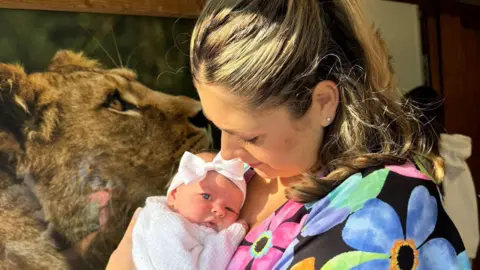 Ami Boyd, who is wearing a black dress with colourful flowers on, is cuddling her daughter Flossie, who is wearing a white knitted cardigan and hair band with a bow. 