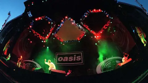 Getty Images Oasis on stage. Green stage lights illuminate them both, with a black backdrop with the Oasis logo on it.
