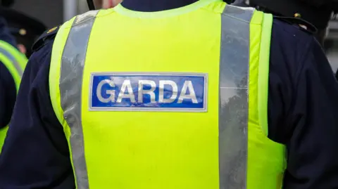 A close-up of the back of a garda officer's uniform.  He is wearing a yellow high-viz vest over a navy uniform. 