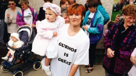 Wearing a 'Gurnos is Great' T-shirt is Gurnos Estate resident Michelle Plummer and daughter Christie, aged 20 months. Michelle has a pixie cut hairstyle which is dyed orange/red and she is holding her daughter in her arms who is wearing all pink. There are other residents behind her and they are standing in front of a pebble-dashed wall.