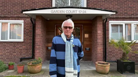 Michael Slade standing outside the block of flats in a blue striped jumper. 