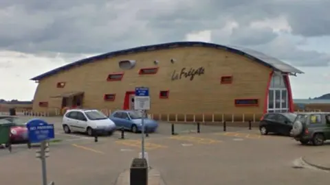 A wooden building looks like an upside down boat, with red window frames. It is located on the seafront. A car park is in front.