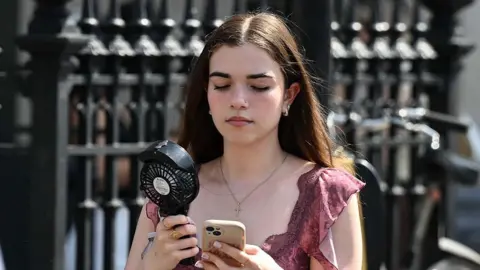 Getty Images A woman looks at her phone while holding a fan near her face