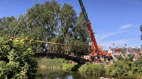 A sunny day with blue sky with a crane in a park area lifting a footbridge, redbrick terraced homes in the distance.