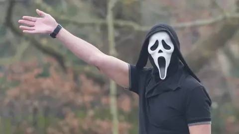 East Anglia News Service A man wearing a mask from the horror film Scream, raising his right arm in a wave. He has a black polo shirt on.