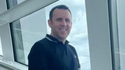 Steven Streeting smiles at the camera. He is clean shaven with short dark hair, and is wearing a black polo shirt. He is standing in front of a large window in a tall building with a city beyond.