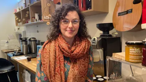 Rosie Hart standing behind the wooden counter at the Roots cafe. She is wearing a blue patterned top with Christmas baubles on it, and an orange and yellow oriental patterned scarf. She has shoulder length dark curly hair and is wearing large glasses. Beside her is a coffee machine and the Perspex display case showing the jars of chutney for sale. There is also an acoustic guitar hung up on the wall to the right.