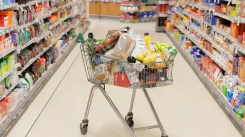 A shopping trolley filled with groceries in the middle of a supermarket aisle