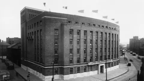 BBC A black and white image of Broadcasting House in Belfast, taken in 1945. Shows the building from the front. There are windows right across the building on six different levels. There are seven flags on the roof.