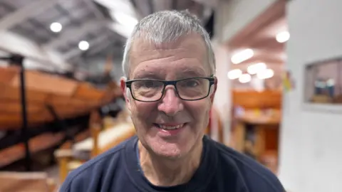 Jo Black/BBC Dave Clemmer stands in front of the ship reconstruction and smiles at the camera. He has short grey hair and wears a navy jumper and glasses. 
