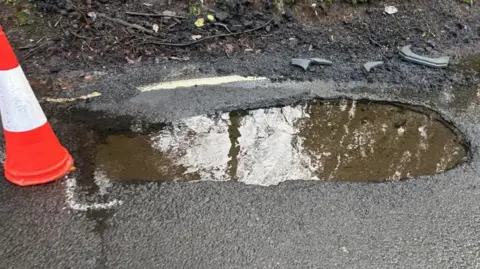 Councillor Craig Monks A traffic cone stands next to a large pothole that is filled with water. There are some white chalk markings around the hole on the road's surface. 