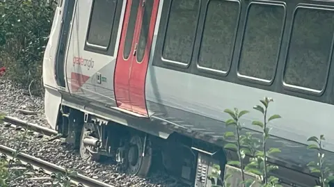 A close-up image of a Greater Anglia railway carriage. Its wheels have left the track and are resting in stones in between the metal lines. The carriage is upright, but at a slight angle.