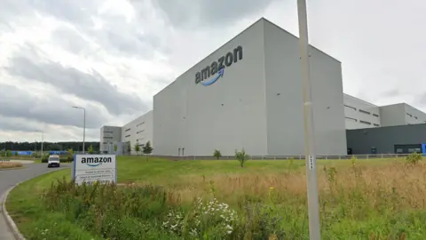 A large grey warehouse with the word "Amazon" in black on its side. Behind it are more grey multi-storey buildings. There is a lawn in front of it with a kerb. The sky is cloudy and grey and there is a sign in front of the building which says: "Amazon"