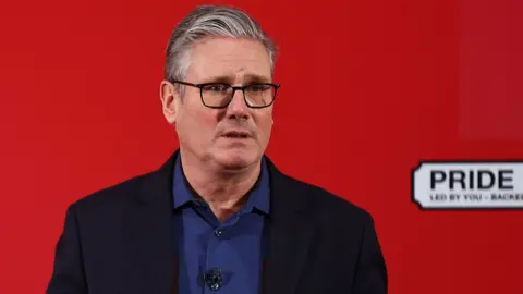 Getty Images Sir Keir Starmer wearing a blue shirt and black suit jacket standing in front of a red background