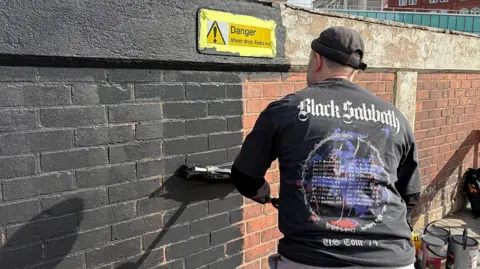 Central BID A man stands facing a brick wall, with his back towards the camera. He is wearing a black Black Sabbath tour-shirt and is painting the wall black. Pots of paint can be seen on the ground next to him.