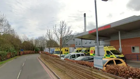 The outside of Hereford's County Hospital. The image is taken from the road side outside the A&E department. The hospital's façade is made up of red brick and grey cladding panels. A small roof with pillars extends out over the car park. Several yellow ambulances are parked beneath it with at least four cars parked in the car park facing away from the hospital.