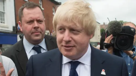 A crowd of people standing around former UK prime minister Boris Johnson. Johnson is wearing a navy blue suit and navy blue tie with a poppy badge. Behind him is Will Walden, who is wearing a black suit and tie. There is also a TV camera operator.