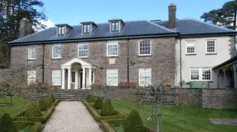 A gravel path through a sculpted Victorian garden leading up to the Romanesque entrance, with white pillars of a large stone mansion flanked by ancient trees and a stone barn 