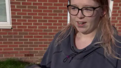 Elouise Evans has long light brown hair and is wearing glasses on her face and a blue sweatshirt. She is standing in front of a redbrick house.