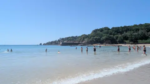A beach on a warm clear sunny day with several people stood in the shallow water near the shoreline. There is a tree-covered hillside and rocky coastline in the background with a stone harbour wall. 