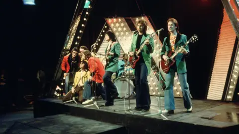 Eight band members stand on a stage playing guitar. Some are wearing green suits and some are wearing red suits. 